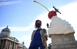 Die Künstlerin Heather Phillipson steht vor einer Skulptur, die einen Sahnewirbel mit einer Kirsche, einer Fliege und einer Drohne zeigt